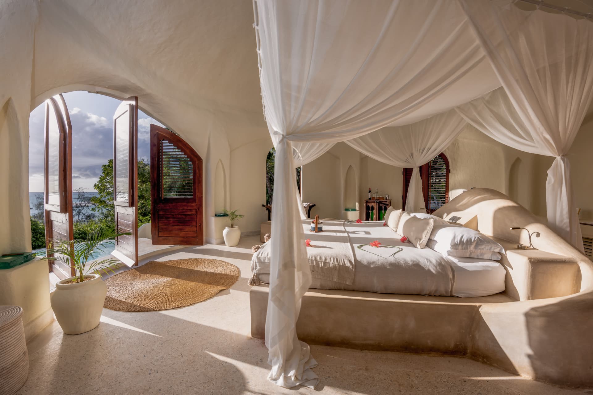 Closed: May 1–31. Kilindi Zanzibar beach villa bedroom, four-poster bed draped in white linens, arched windows open to the Indian Ocean