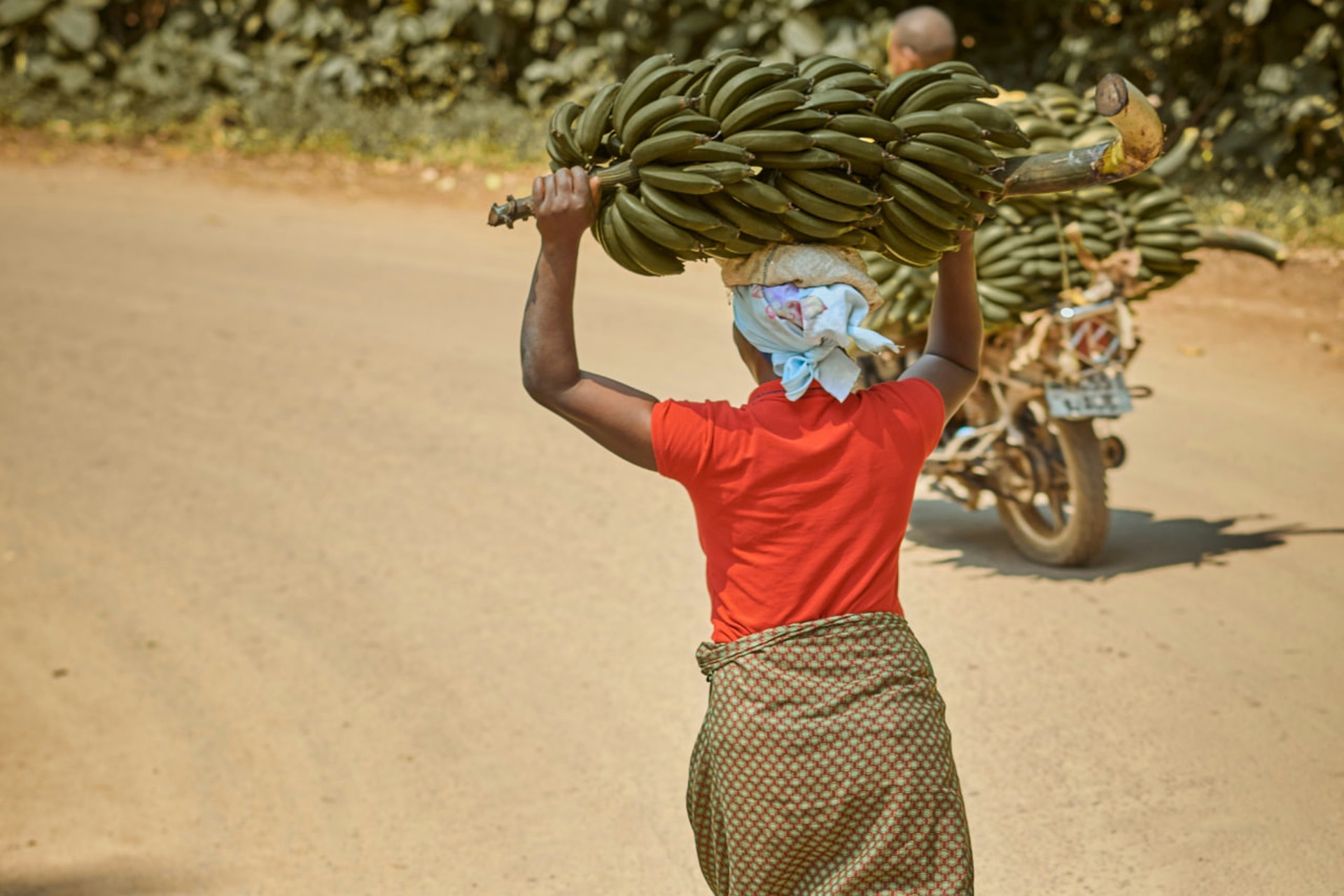 A woman walking along a dusty road in northern Tanzania, carrying a large bunch of bananas on her head