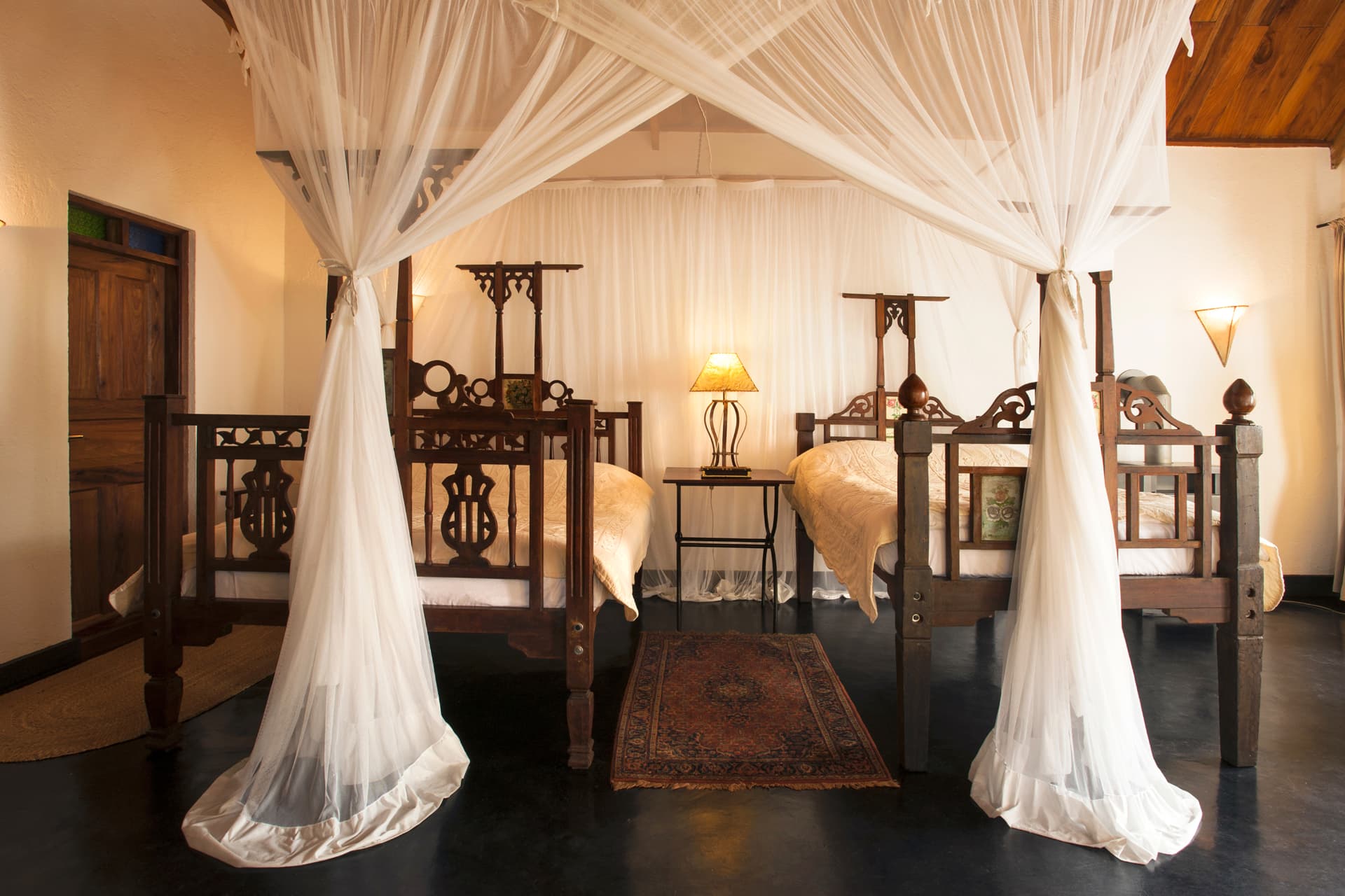 The Plantation Lodge twin room with two carved antique wooden beds under white mosquito nets, warm lamplight and a Persian rug near Karatu